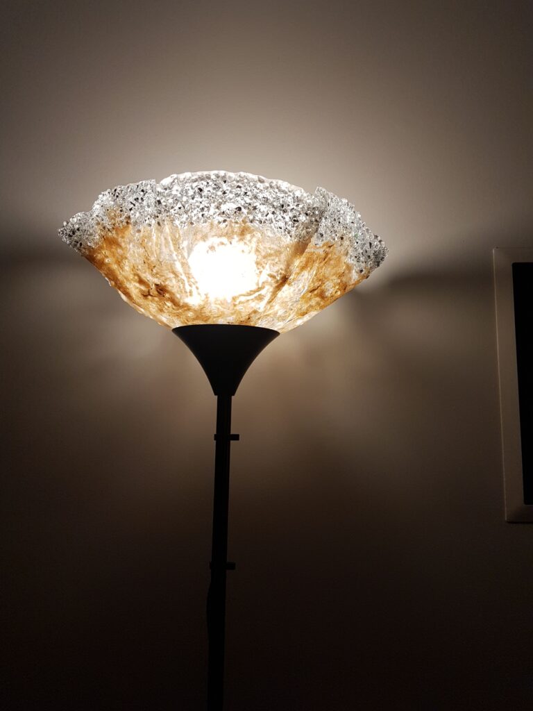 Resin Lampshade An illuminated floor lamp with a textured, shimmering resin lampshade casting a warm glow upwards, creating ambient light in a room.